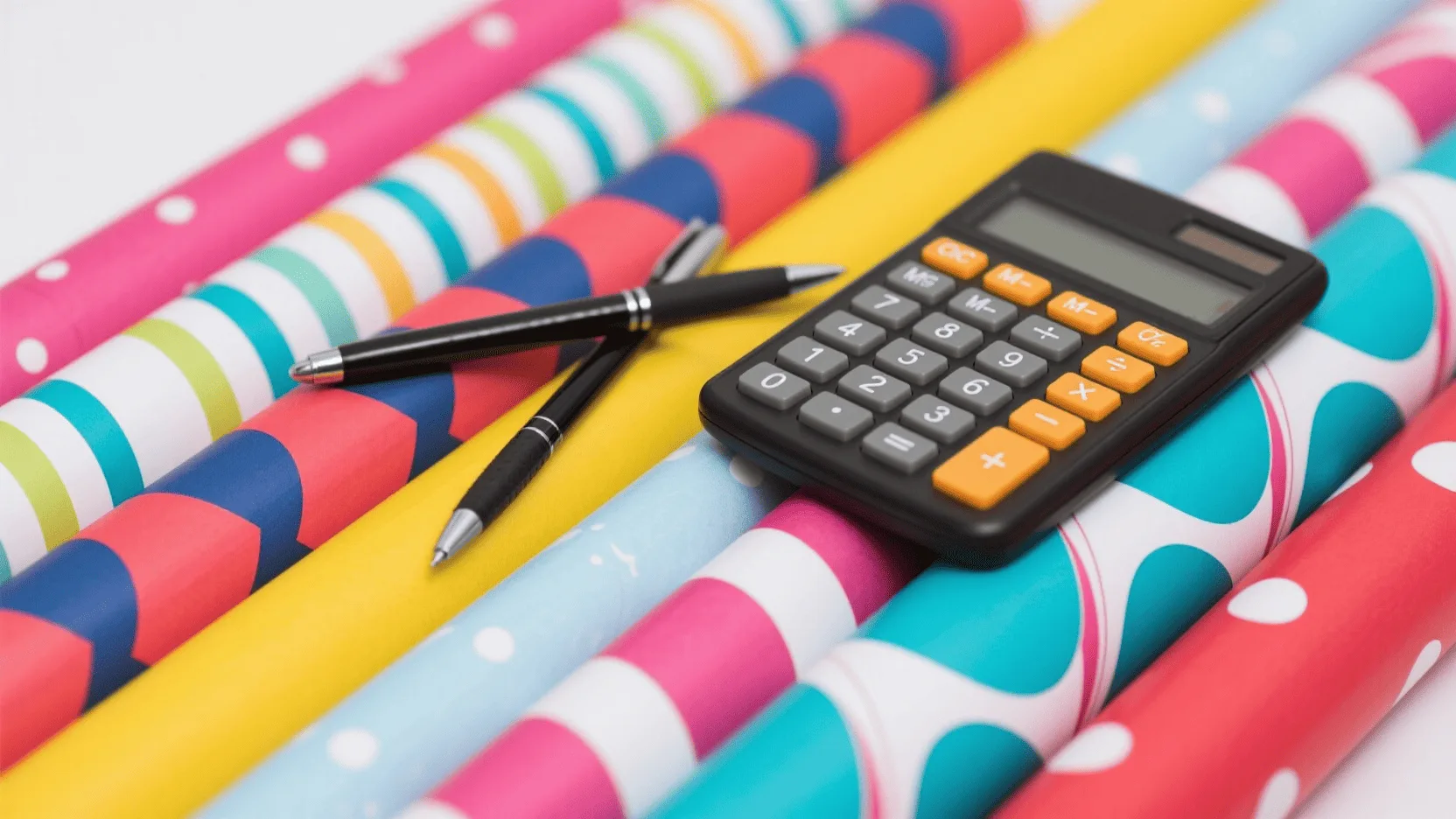 A calculator and pen resting on top of several rolls of colorful wholesale wrapping paper.