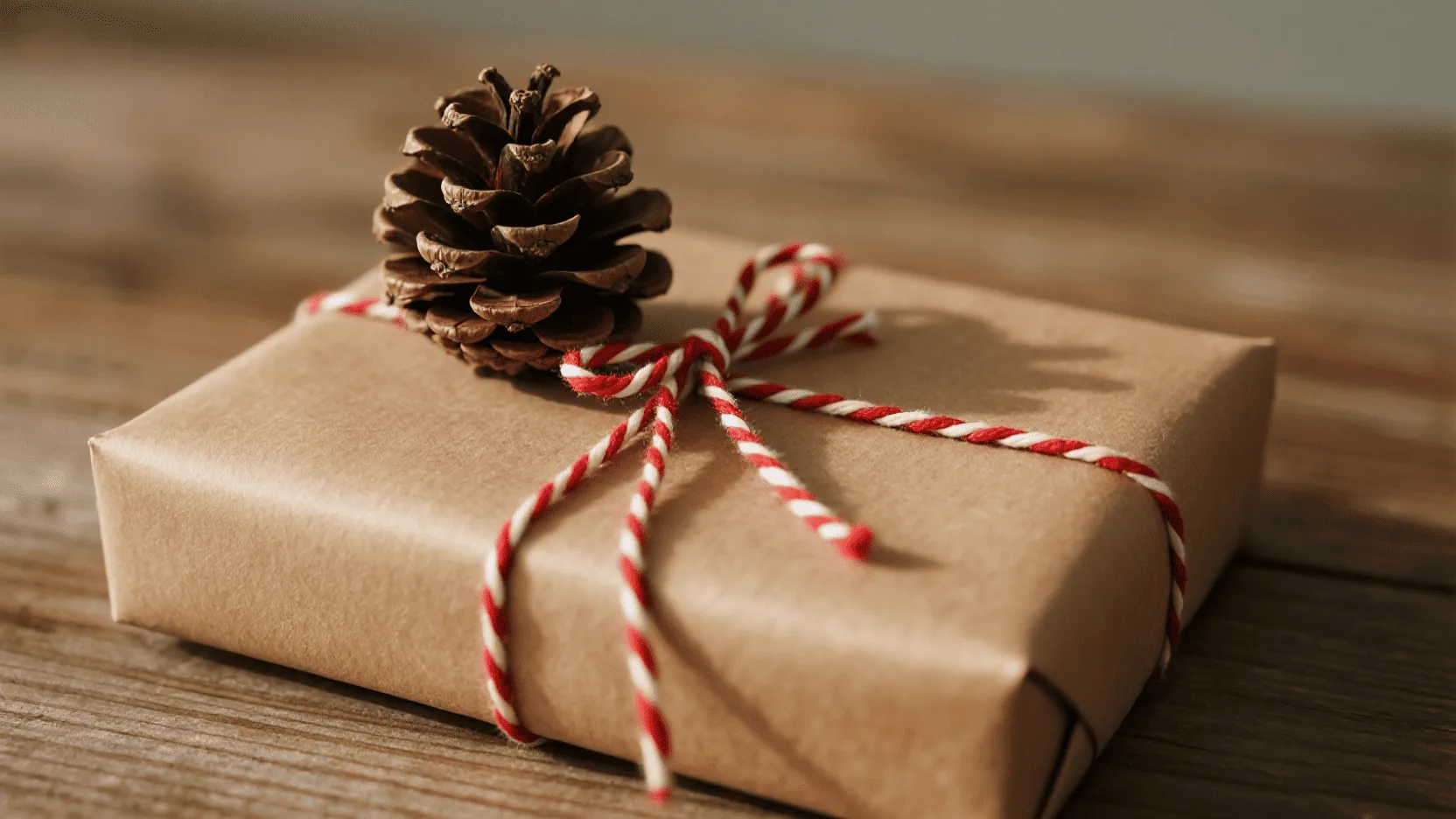 A beautifully wrapped gift in brown kraft paper, decorated with red and white twine and a small pinecone.