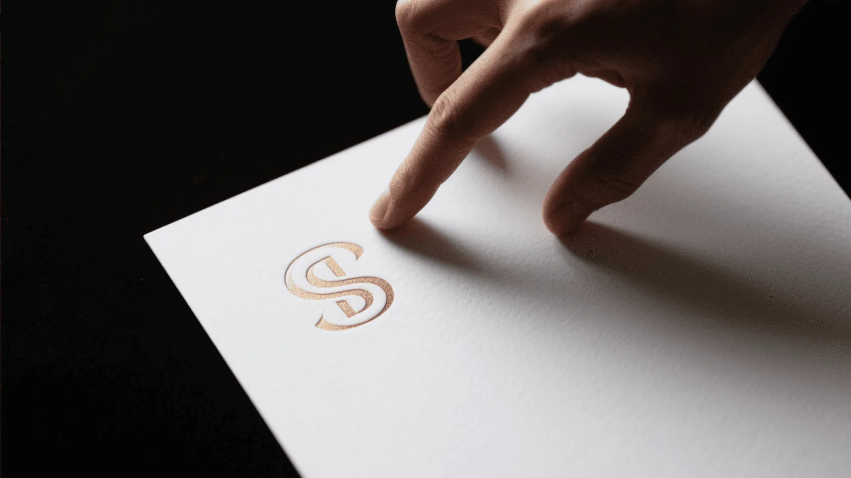 A hand touching a white paper with an elegant embossed logo, showing the raised texture.
