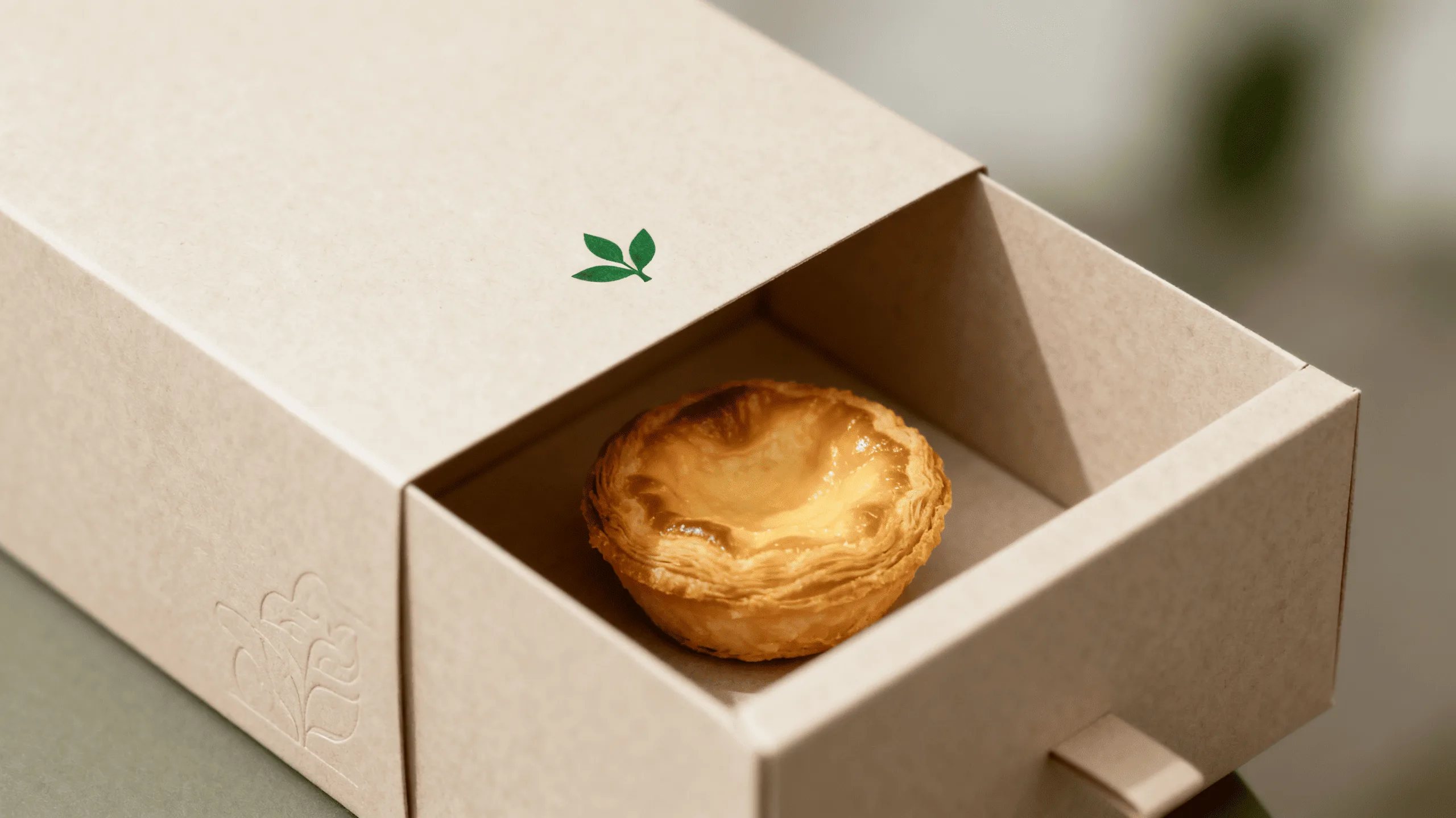 A beautifully crafted, minimalist paper box holding a delicate pastry, with a small green leaf emblem on it.