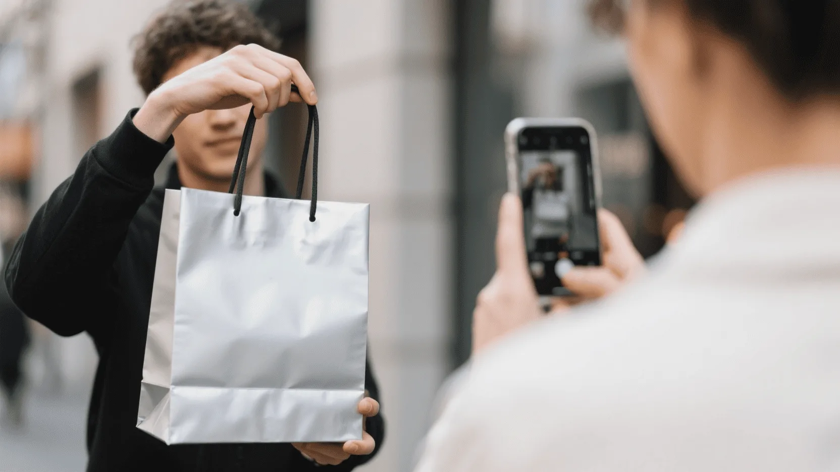 A person holding a stylish silver paper bag, taking a photo of it with their smartphone for social media.